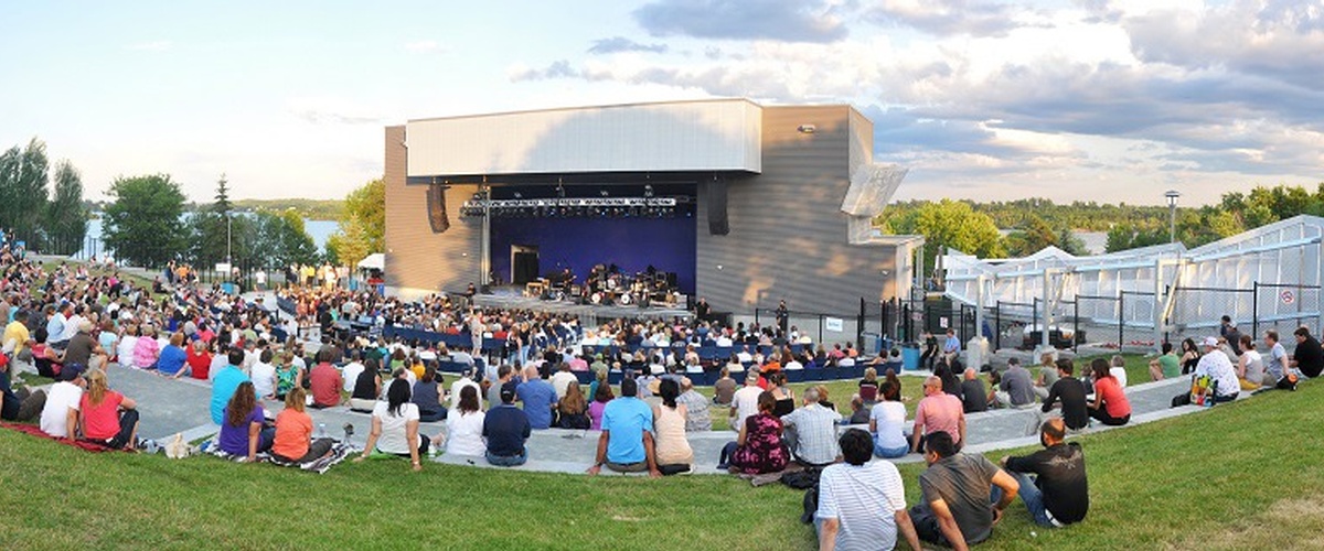 Outdoor amphitheatre by the lake, on a sunny afternoon. People fill the seats as they watch the stage.